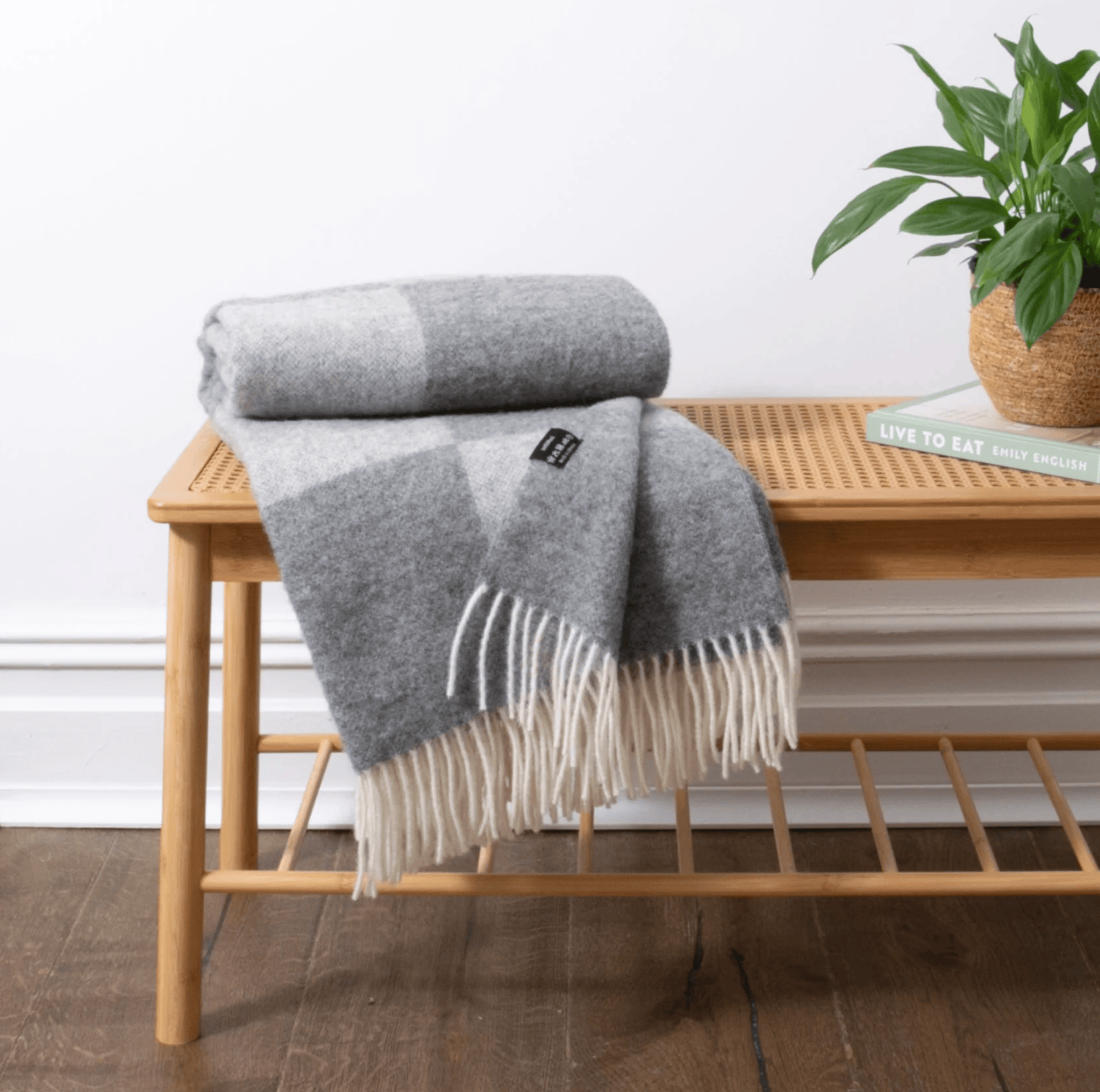 Grey checkered blanket with fringes on a wooden bench, plant and book in the background.