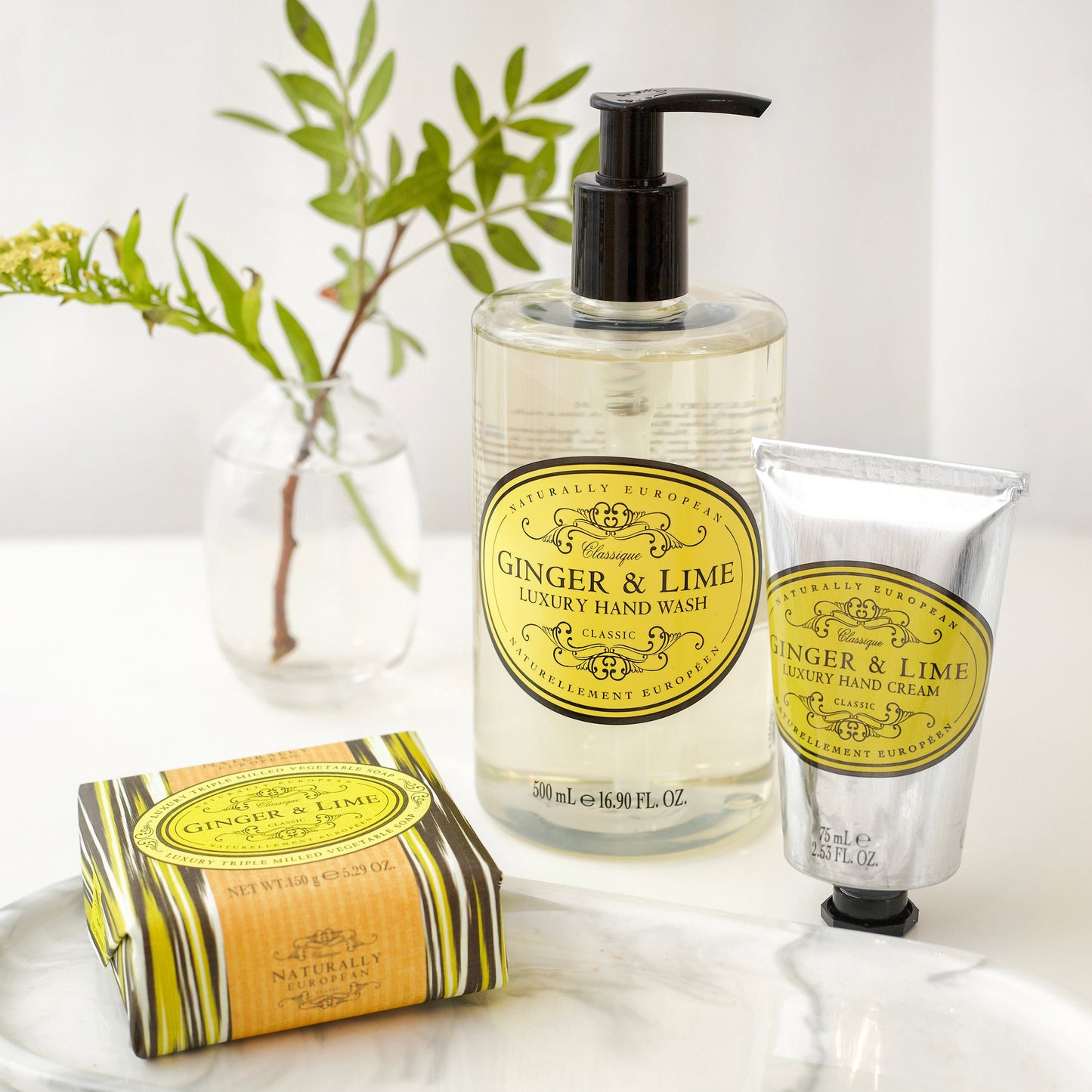 Somerset Toiletries Ginger & Lime luxury hand wash, cream, and soap on a white surface with greenery in the background.