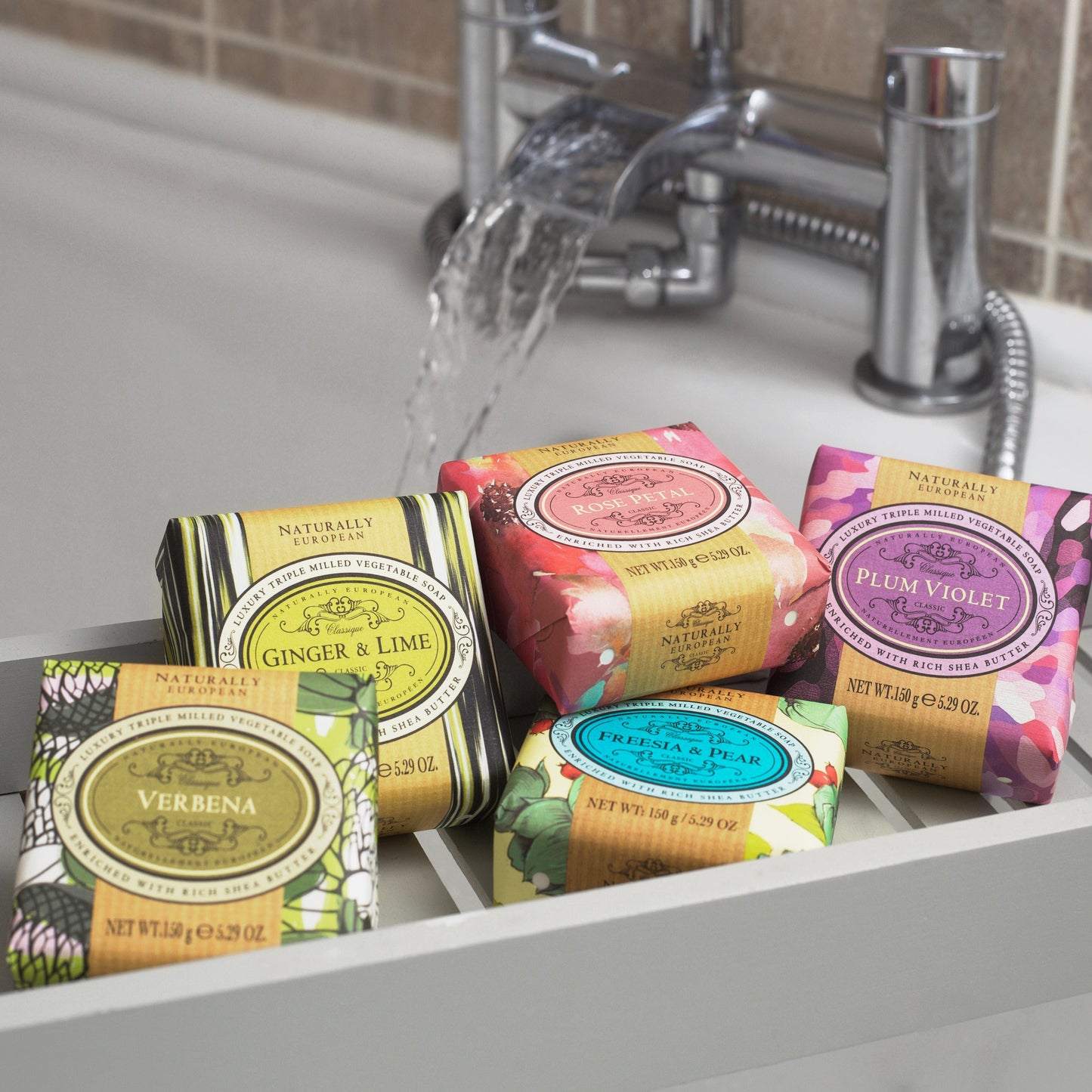 Five Somerset Toiletries Naturally European soap bars with colourful packaging on a sink, water flowing in the background