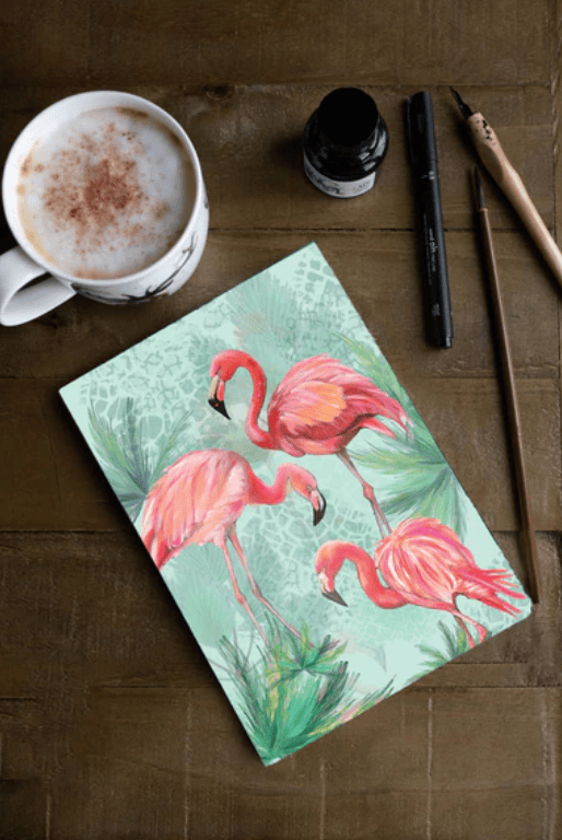 Notebook with flamingo design on a wooden surface with a cup of coffee and writing utensils.