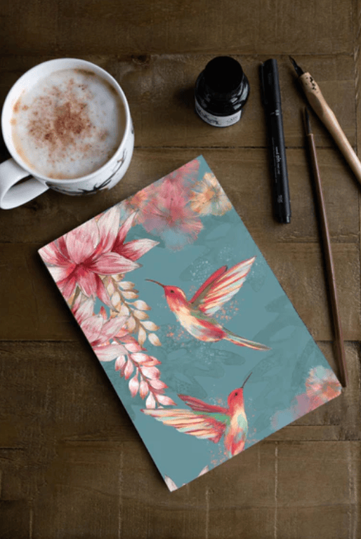 Notebook with floral and hummingbird design on a wooden surface with a cup of coffee and writing utensils.