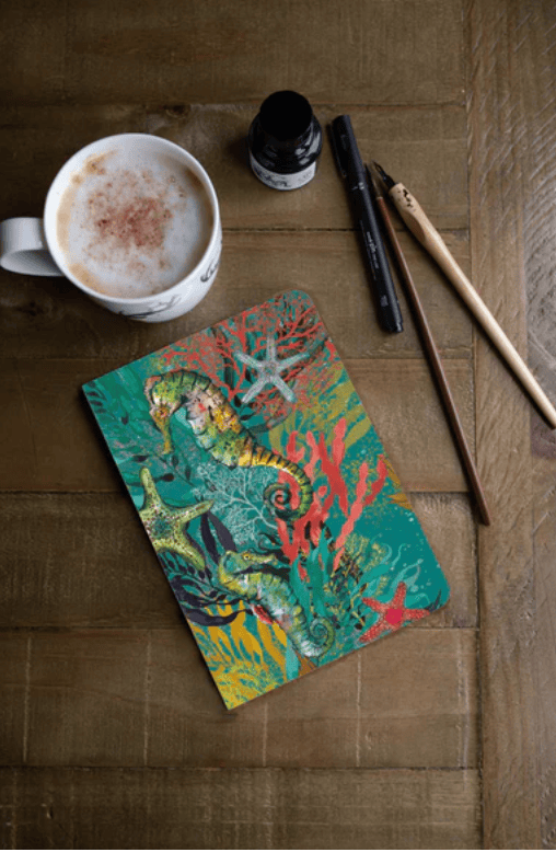Colorful notebook with seahorse design on a wooden surface with a cup of coffee and stationery items.