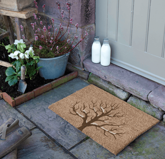 Doormat with tree design on a stone pathway with plants and milk bottles.