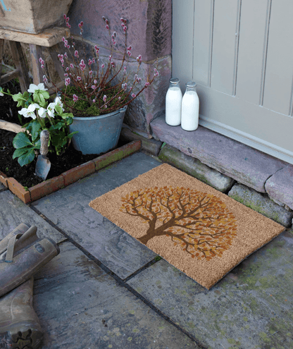 Doormat with tree design on a stone pathway, with plants and bottles in the background.