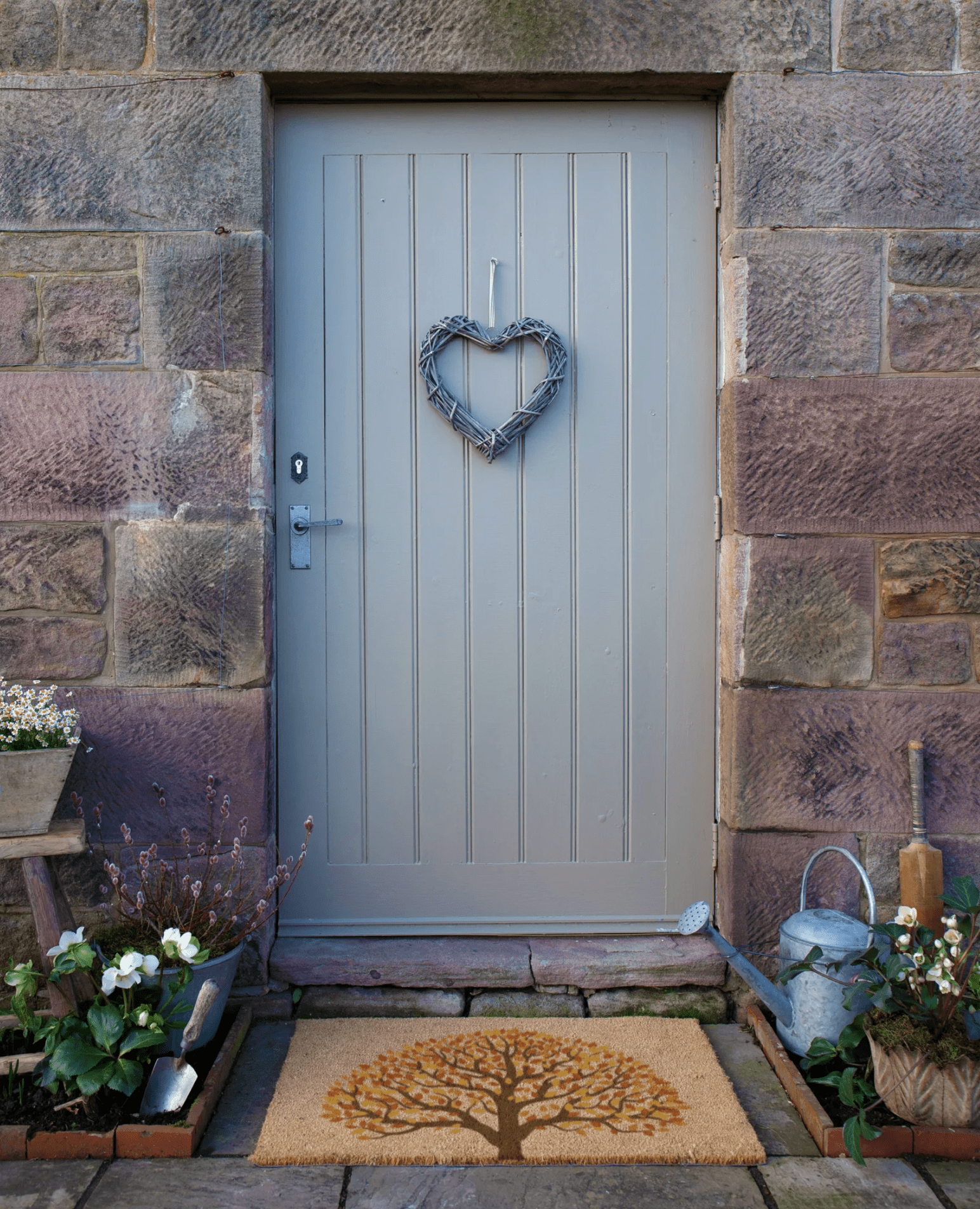 Gray front door with a heart-shaped wreath on a stone wall, surrounded by plants and decor.