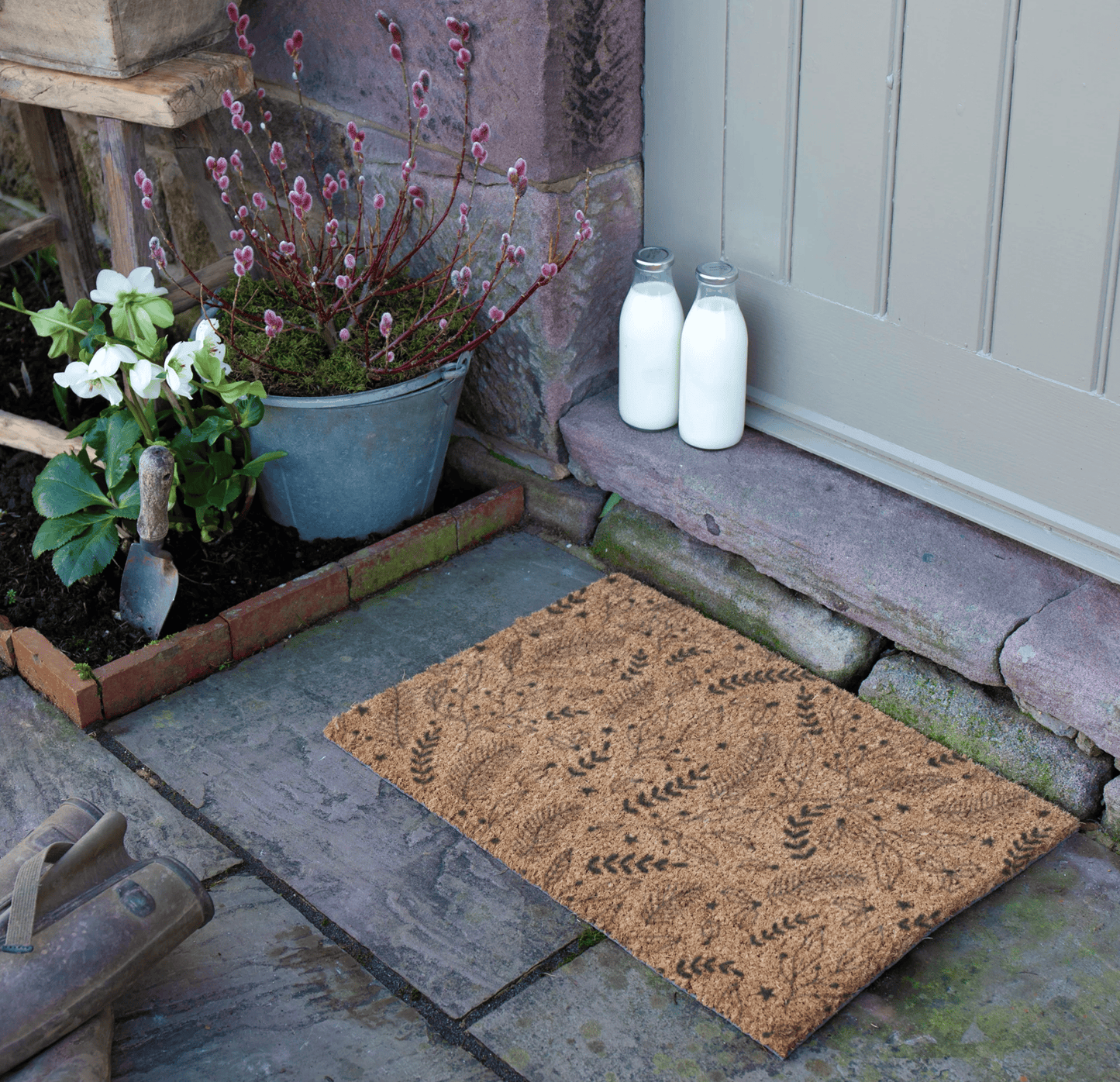 Doormat with leaf pattern on a stone pathway, with plants and milk bottles in the background.