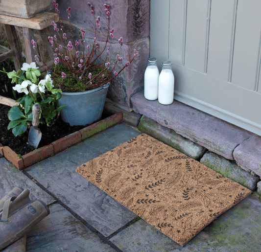 Doormat with leaf pattern on a stone pathway, with plants and milk bottles in the background.