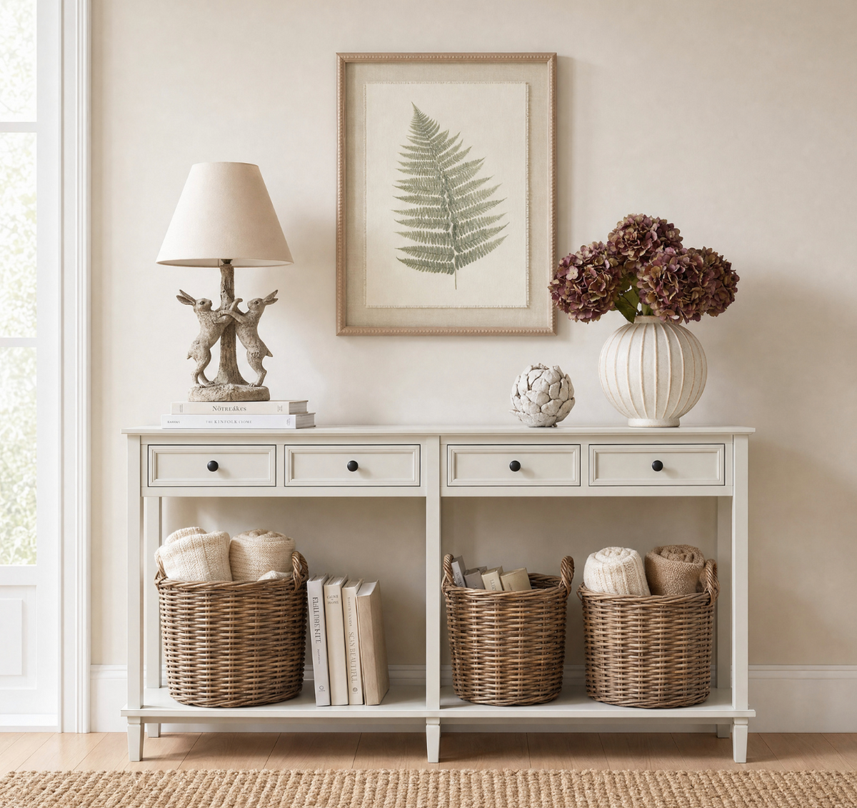 A white console table styled in a bright, neutral-toned room with a framed fern print, a deer lamp, a vase of purple hydrangeas, and three wicker baskets on the lower shelf.