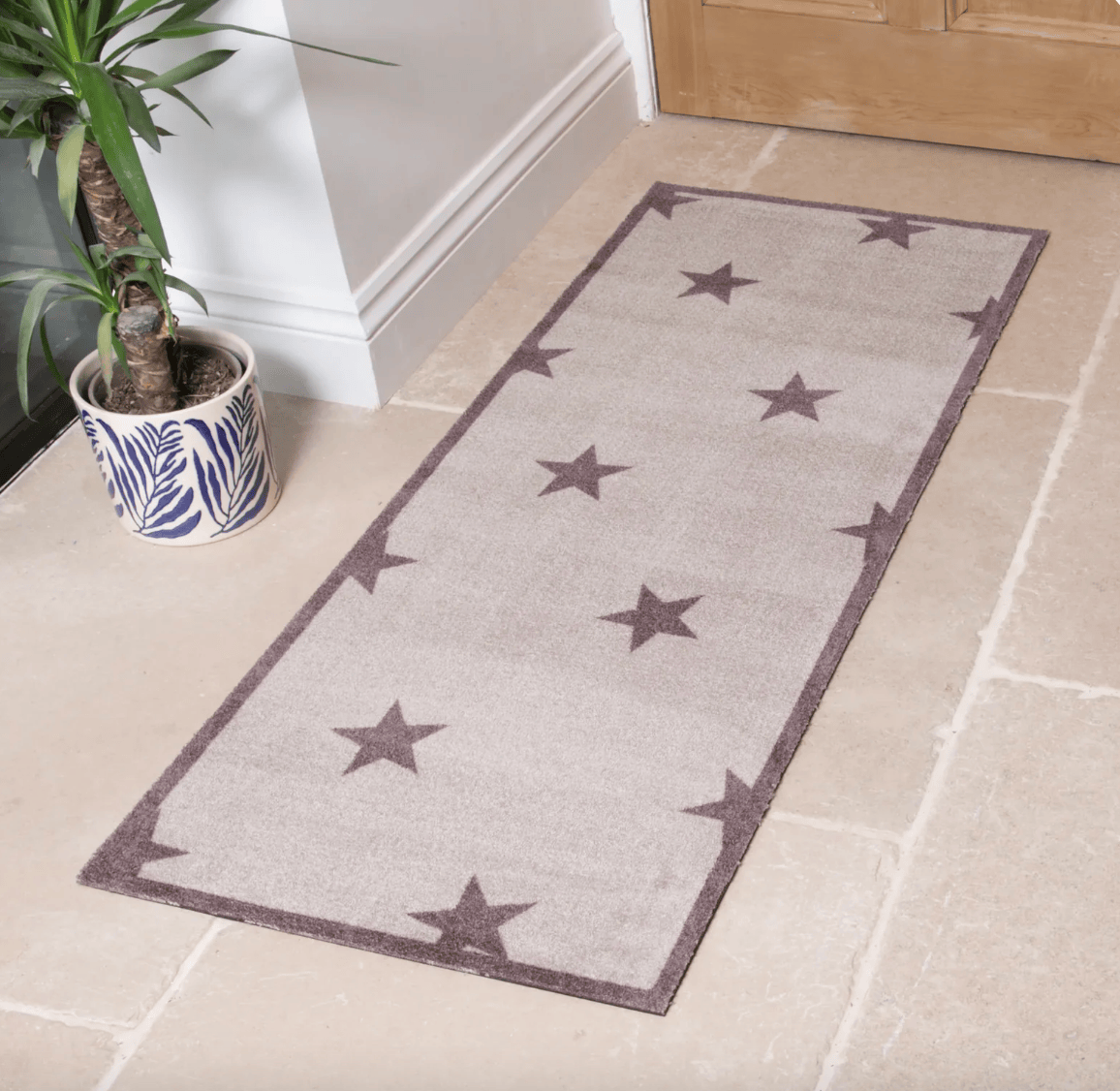 A long rectangular entryway rug with a taupe star pattern and border, shown on a light stone floor next to a potted plant and a wooden door.