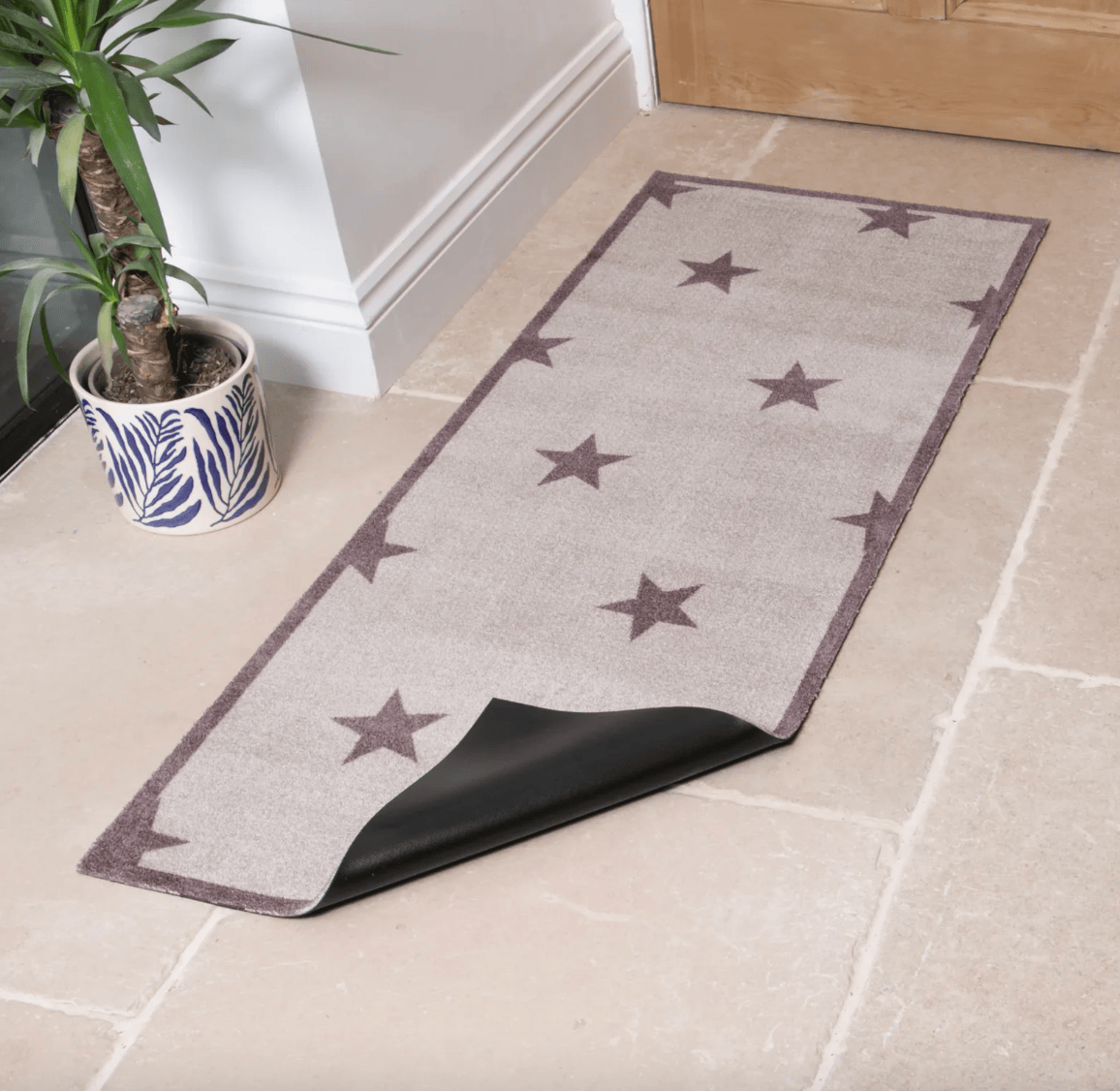 A rectangular entryway rug with a beige base, dark brown star pattern, and a black peel-up backing, shown on a light stone floor next to a potted plant.