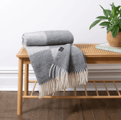 Grey checkered blanket with fringes on a wooden bench, plant and book in the background.
