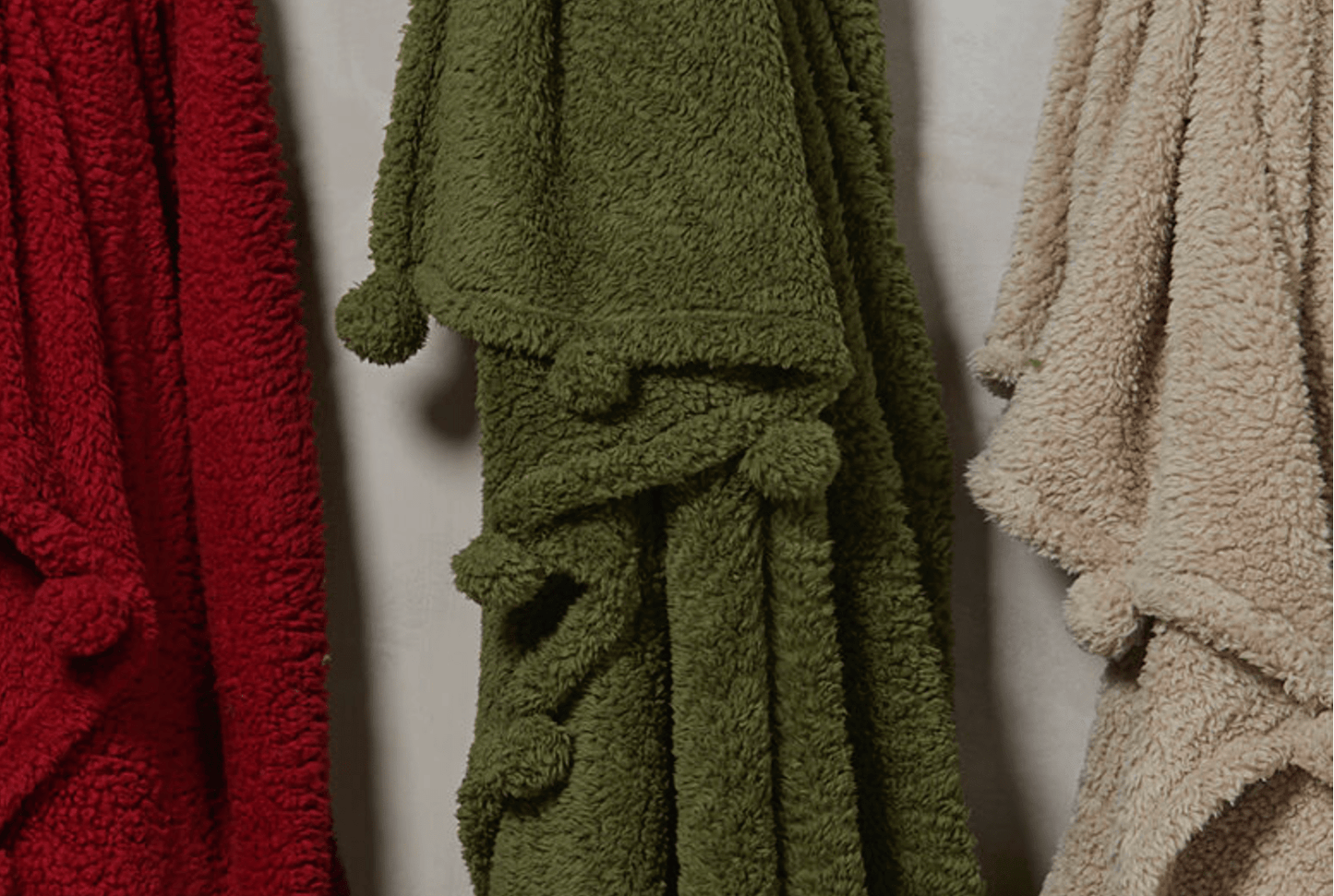 Three plush bathrobes in red, green, and beige hanging against a neutral background.