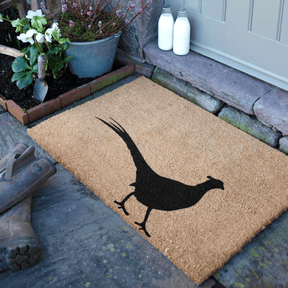 Doormat with a black bird design on a stone pathway, with plants and milk bottles in the background.