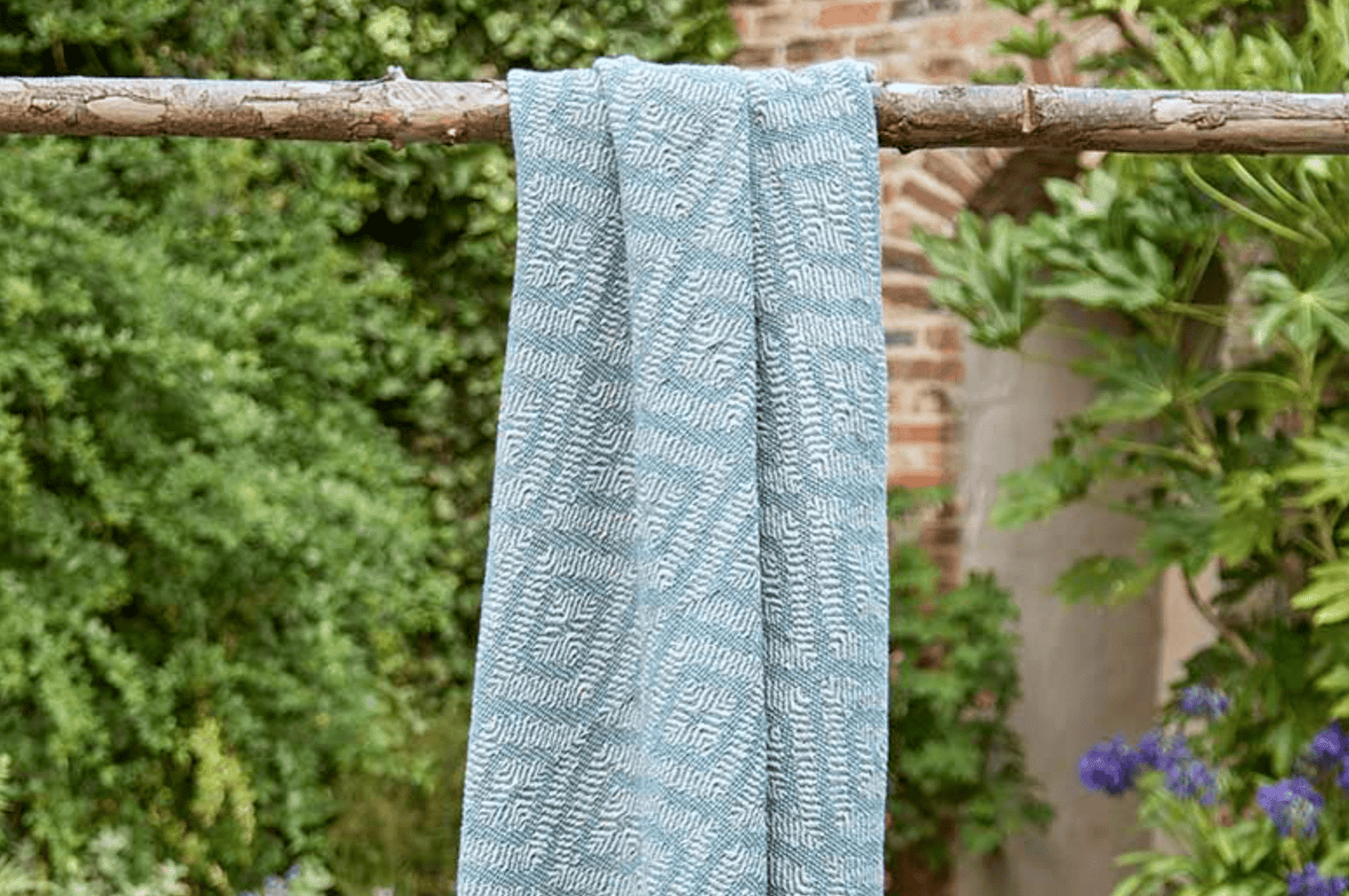 Blue textured towel hanging on a wooden stick with greenery and a building in the background