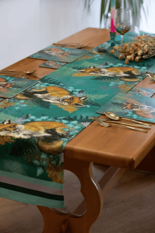 A styled dining table featuring a green table runner and matching placemats with a detailed print of foxes in a forest, set with gold cutlery on a wooden table.
