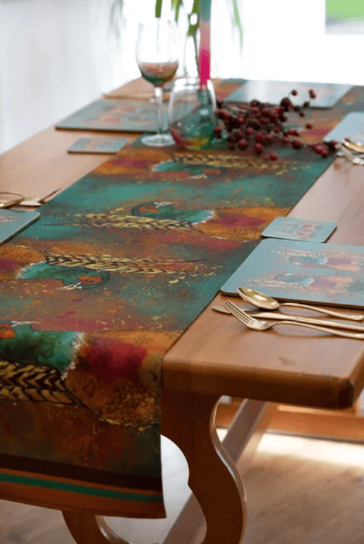 A colorful abstract-patterned table runner and matching placemats styled on a wooden dining table with gold cutlery, wine glasses, and berries.