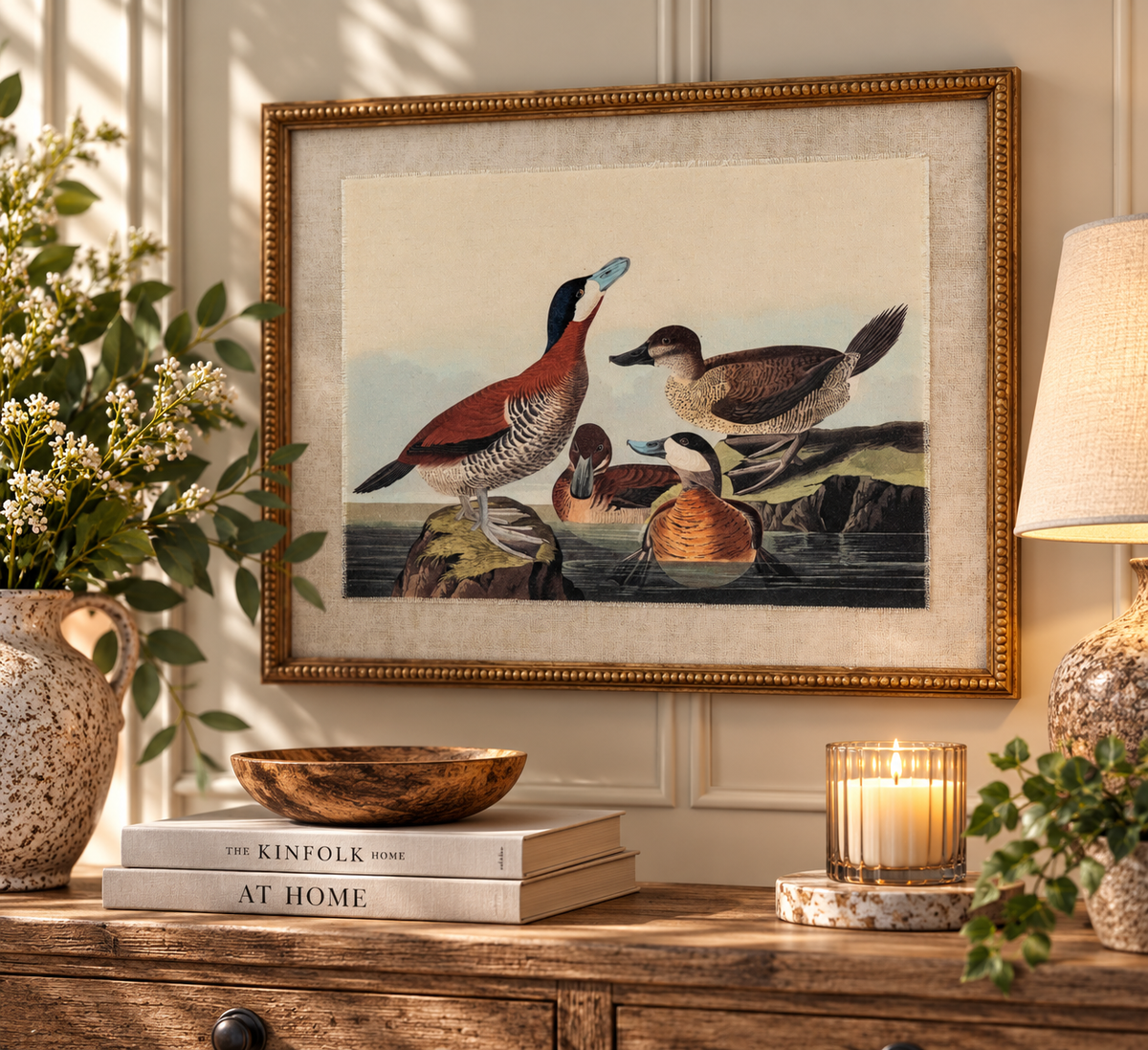 A framed vintage-style duck painting hangs on a cream paneled wall above a wooden console table styled with books, a bowl, a candle, and potted plants. The frame is gold-toned, and the artwork shows several ducks and waterfowl in natural colors.