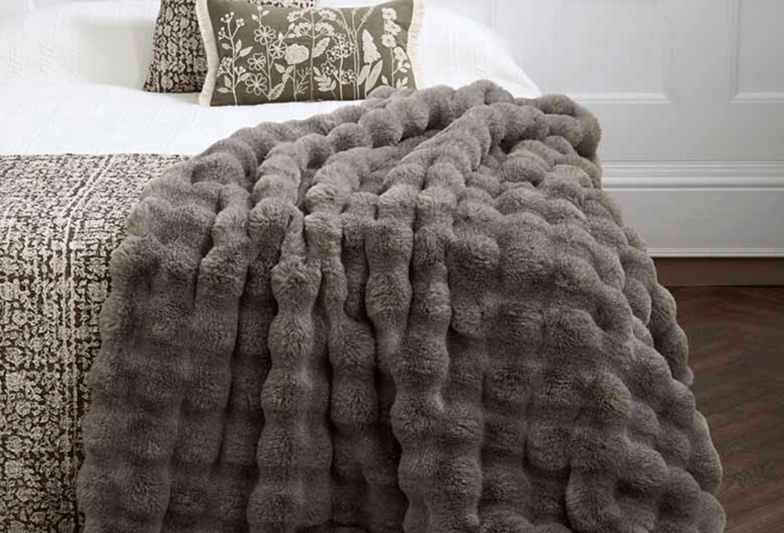 Gray textured blanket draped over a bed with pillows in a bedroom setting.