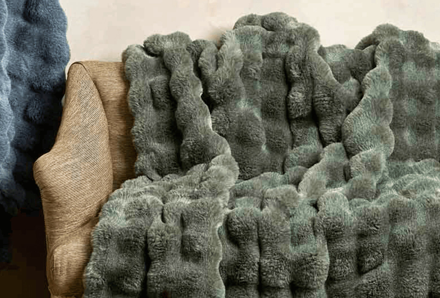 A close-up of a plush, chunky knit throw blanket in a muted sage green color draped over a beige upholstered armchair.