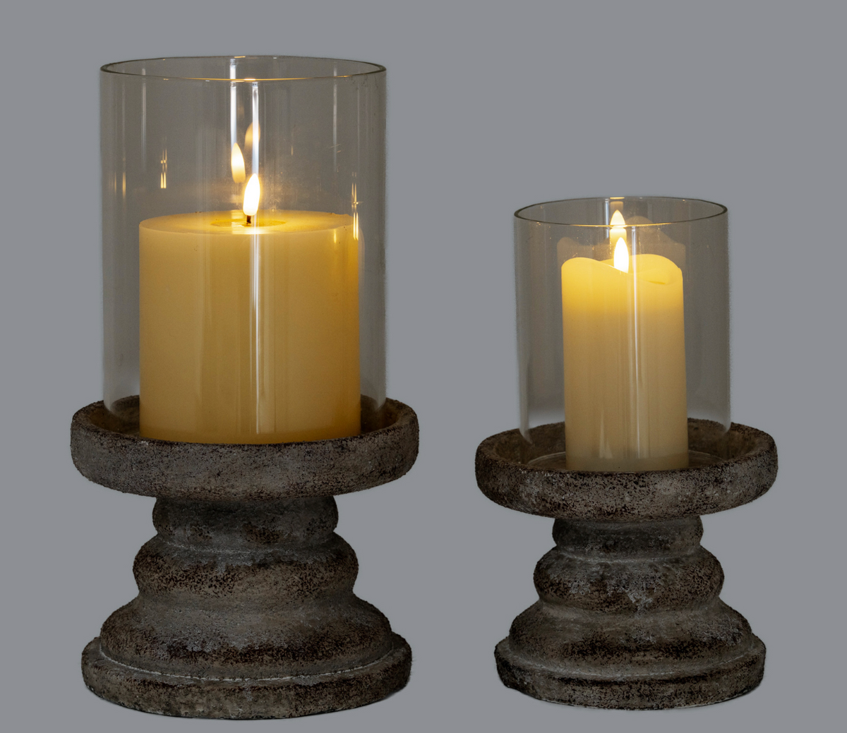 Two lit pillar candles in clear glass cylinder holders on distressed dark metal candle stands, shown against a solid gray background.
