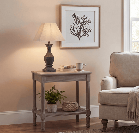 A rustic gray wooden end table styled in a cozy living room setting, holding a table lamp, books, a mug, a small potted fern, and a woven basket. The table is positioned against a beige wall under a framed print, next to a beige armchair.