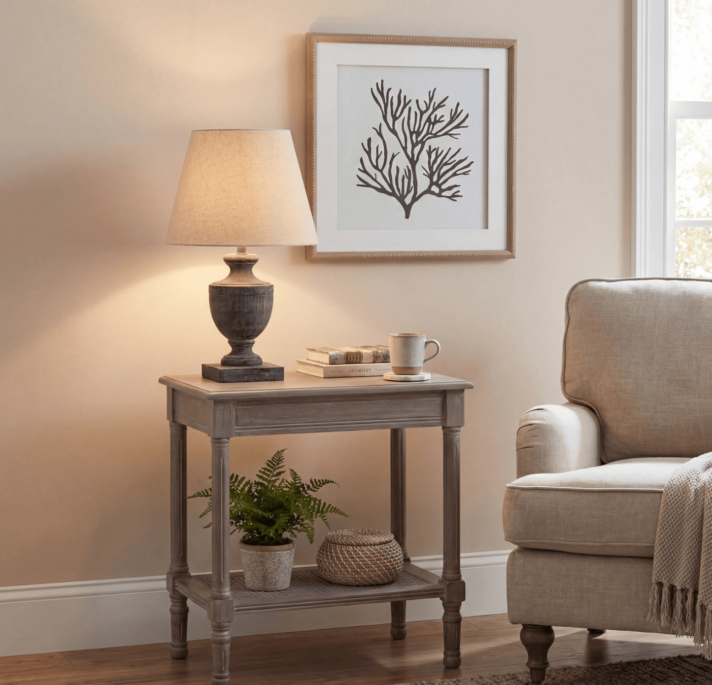 A rustic gray wooden two-tier end table styled in a cozy living room setting, holding a lit table lamp, a stack of books, and a small mug. A small potted fern and a woven basket sit on the lower shelf.
