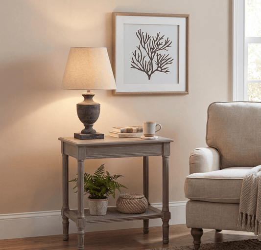 A rustic gray wooden two-tier end table styled in a cozy living room setting, holding a lit table lamp, a stack of books, and a small mug. A small potted fern and a woven basket sit on the lower shelf.