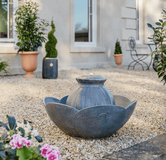 A gray stone lotus-shaped fountain with a central rounded pool, placed outdoors on a bed of light gravel in a garden courtyard with potted plants and a building facade in the background.