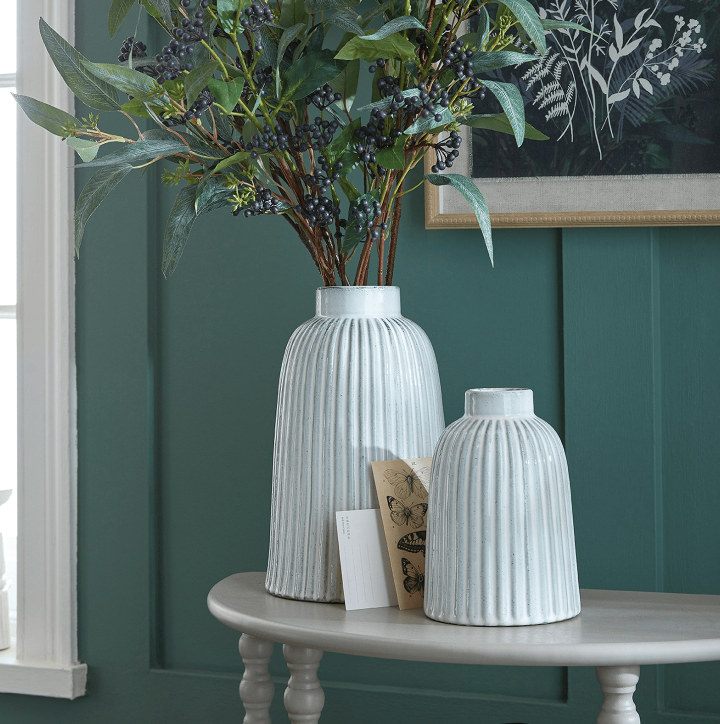 Two white textured vases on a table against a green wall with a framed picture.