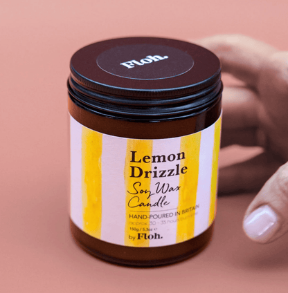 Jar of Lemon Drizzle soy wax candle with a hand holding it on a pink background