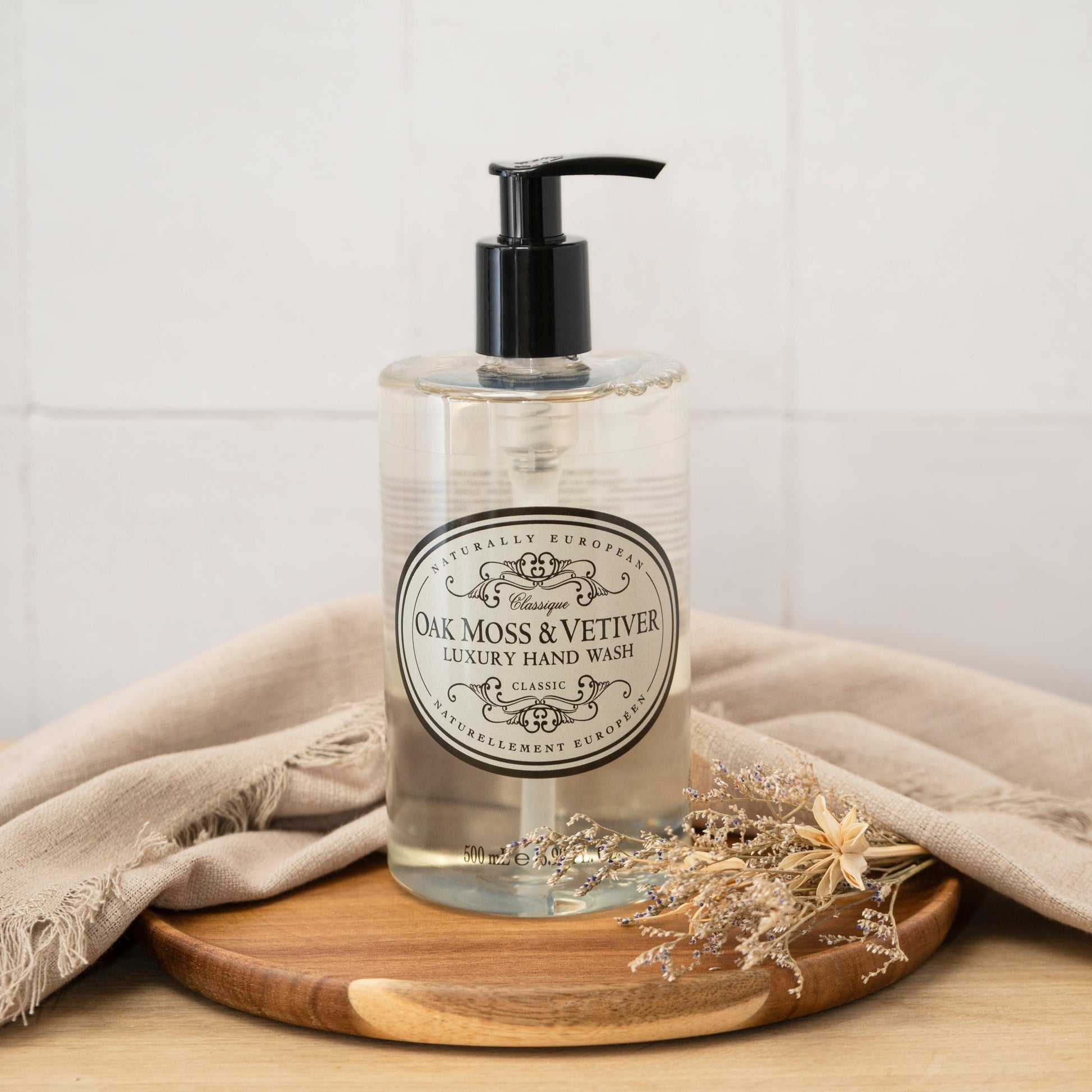 Bottle of Oak Moss & Vetiver Luxury Hand Wash on a wooden tray with a beige cloth and dried flowers.