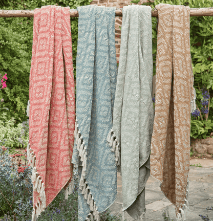 Four patterned scarves in pink, blue, green, and beige hanging on a wooden rod outdoors.