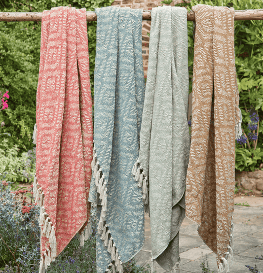Four patterned scarves in pink, blue, green, and beige hanging on a wooden rod outdoors.