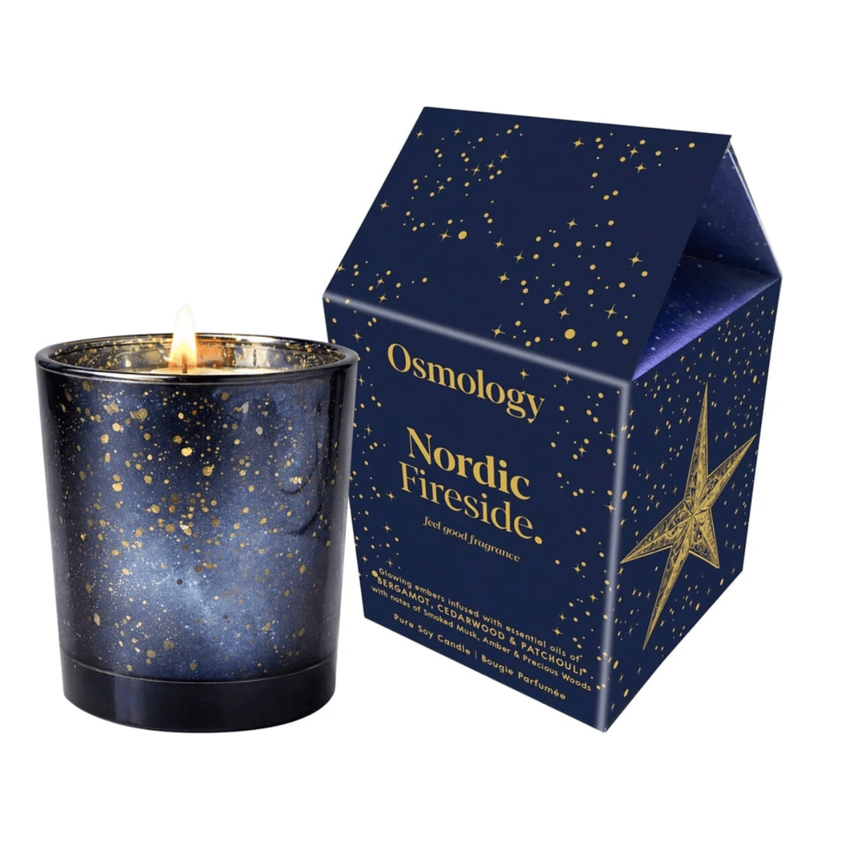 A lit Osmology Nordic Fireside scented candle in a dark blue galaxy-patterned glass jar, shown next to its matching house-shaped packaging box with gold star and speckle details on a white background.