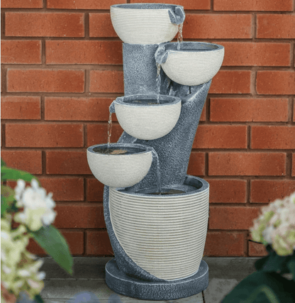 A four-tier outdoor water fountain with a textured gray stone-like body and cream-colored ribbed bowls, set against a red brick wall with blurred flowers in the foreground.