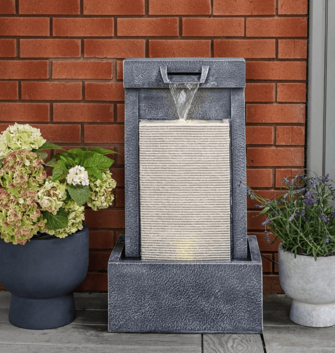 A rectangular outdoor water fountain with a textured gray stone-look frame and a ribbed light beige waterfall panel, shown on a patio against a red brick wall with potted plants on both sides.