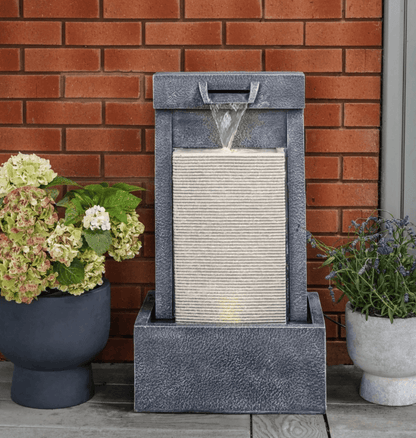 A rectangular outdoor water fountain with a textured gray stone-look frame and a ribbed light beige waterfall panel, shown on a patio against a red brick wall with potted plants on both sides.