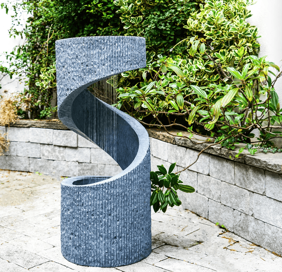 A modern spiral outdoor water feature with a textured gray stone-like finish, set on a paved patio with a stone wall and green foliage in the background.