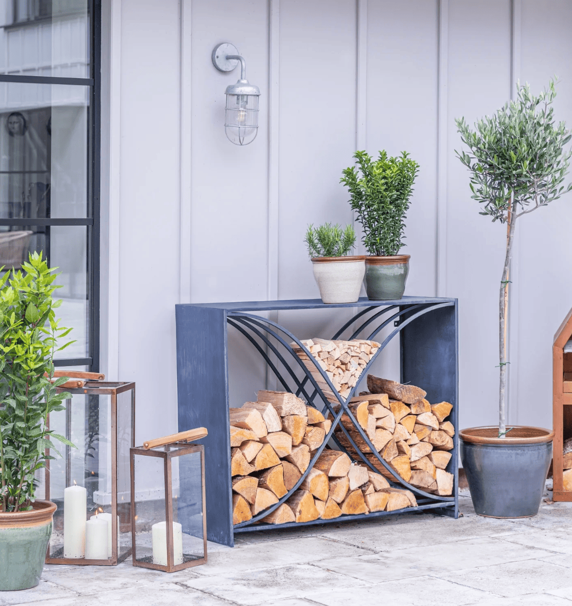 Outdoor firewood storage rack with stacked logs, potted plants, and lanterns on a patio.