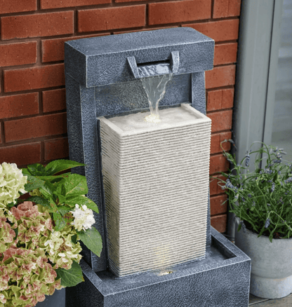 A rectangular outdoor water fountain with a dark gray textured stone finish and a lighter beige stone waterfall chamber, set against a red brick wall with potted plants on either side.