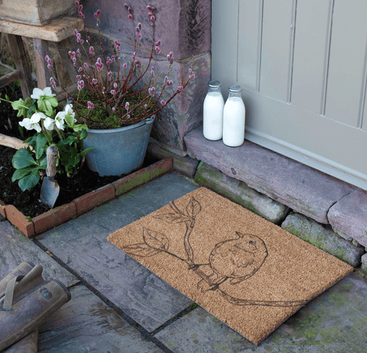 Doormat with a design on a stone pathway next to a door, with plants and milk bottles in the background.