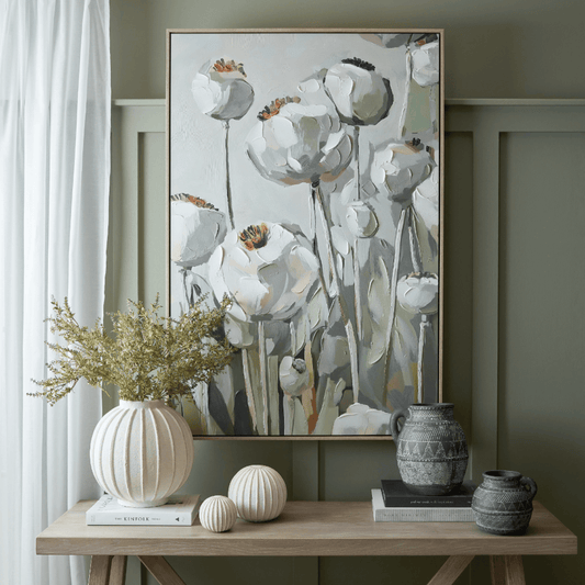 A framed vertical canvas painting of white poppy flowers with muted green and gray stems, displayed on a wooden console table in a styled room setting with decorative vases and books.