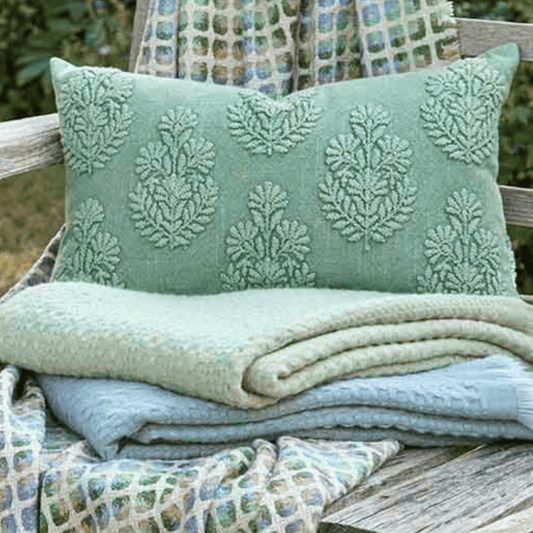 Green textured pillow and blanket on a wooden bench with a patterned fabric background.
