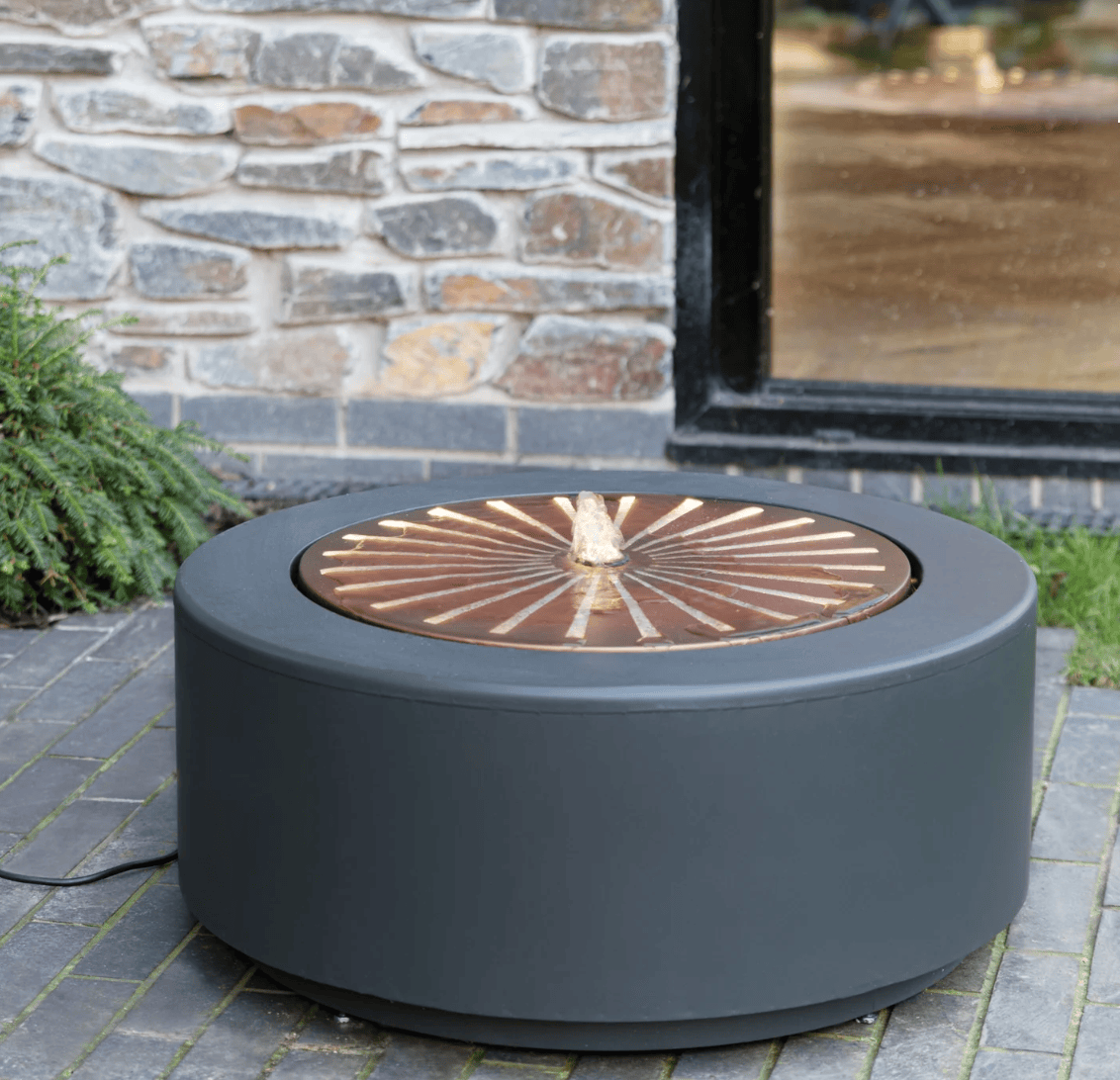 A round outdoor fire pit table with a matte dark gray body and a copper-toned circular insert with a central flame-shaped detail, shown on a stone patio in front of a stone wall and window.