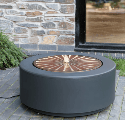 A round outdoor fire pit table with a matte dark gray body and a copper-toned circular insert with a central flame-shaped detail, shown on a stone patio in front of a stone wall and window.