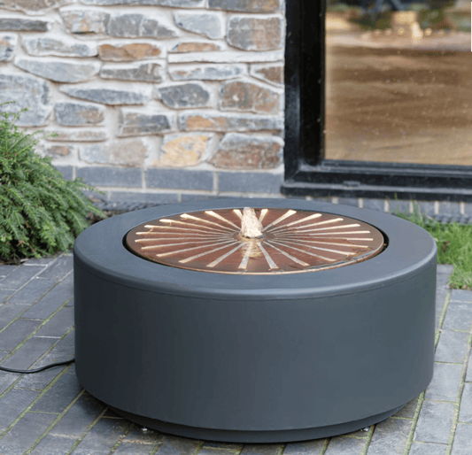 A round outdoor fire pit table with a matte dark gray body and a copper-toned circular insert with a central flame-shaped detail, shown on a stone patio in front of a stone wall and window.