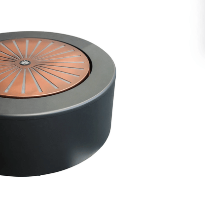A close-up of a modern cylindrical table or stool with a matte black body and a circular copper-toned top featuring a sunburst pattern.