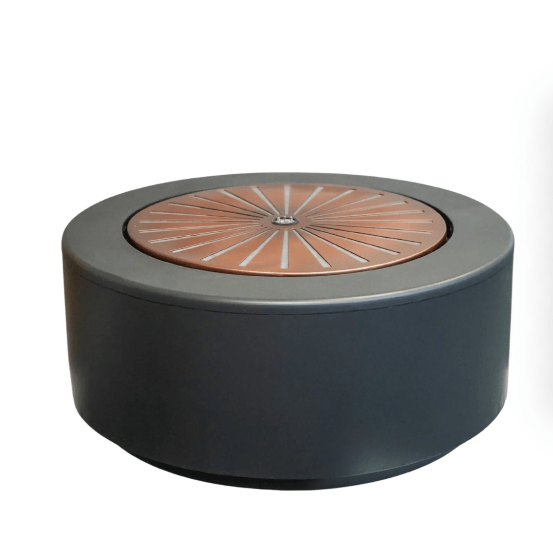 A round outdoor fire pit table with a matte black cylindrical body and a copper-toned metal lid featuring a sunburst pattern.