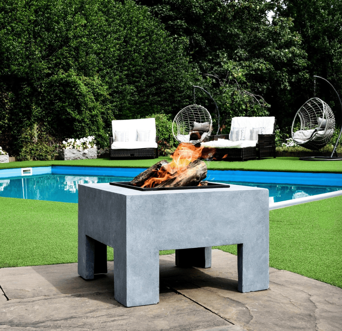A square concrete fire pit table with a black firebox and active flames, situated outdoors on a stone patio next to a swimming pool with green grass and lounge chairs in the background.
