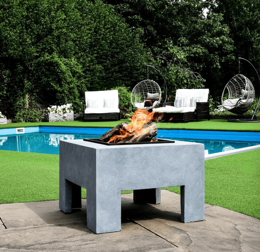 A square concrete fire pit table with a black firebox and active flames, situated outdoors on a stone patio next to a swimming pool with green grass and lounge chairs in the background.